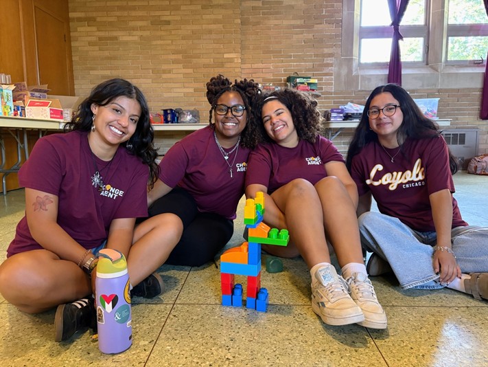 Four girls in maroon shirts sit on the floor, playing with a toy giraffe between them.